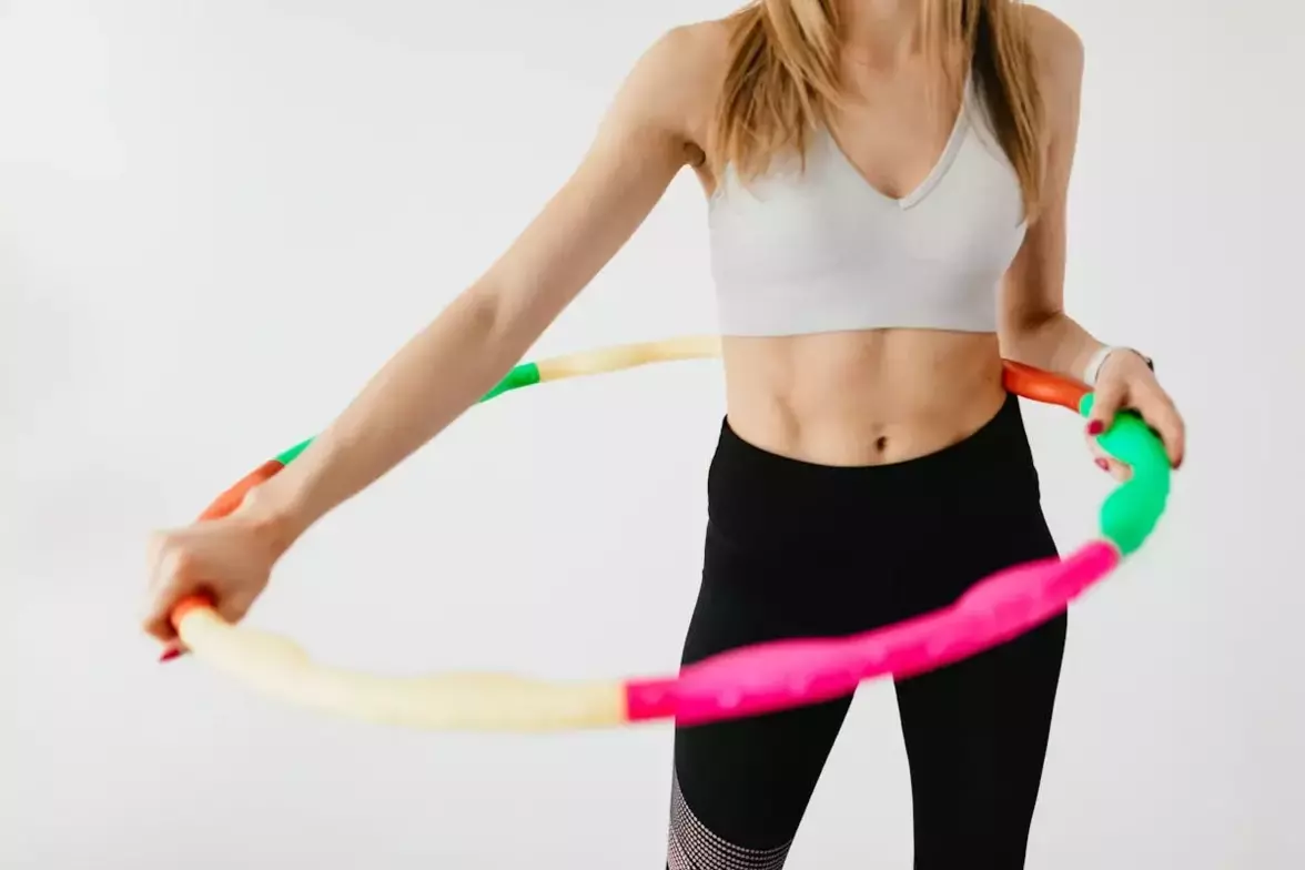 Young woman in athletic wear holding a colorful hula hoop, demonstrating an exercise for weight loss, against a plain background.